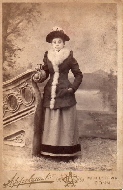 Cabinet card by the Appelquest studio in MIddletown, CT. Shows a full-length view of a woman standing by a studio set. The woman is wearing a fur-trimmed jacket. (Courtesy McIntyre-Culy collection).