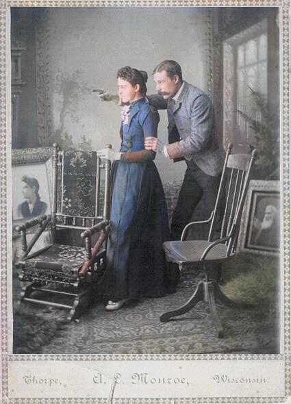 Cabinet card photo of man and woman standing in a room surroundd by large photos and desk chairs. Colorized - the woman is in a blue skirt and jacket. Photo by A. L. Monroe studio, Thorpe, Wisconsin. Posted by user on ancestry.com and said to be of A.L. Monroe and his wife, Allie Monroe.