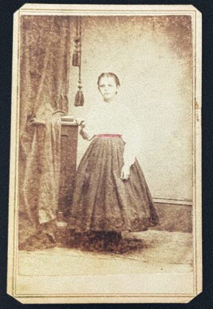 Black and white photo of a young girl wearing a hand-tinted colorful belt. Taken in the Carmel,  NY studio of J. H. Folsom, circa 1860s